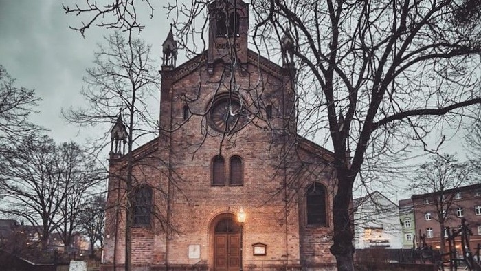 Historische Kirche mit beleuchtetem Uhrturm auf einem nassen Kopfsteinpflasterplatz.