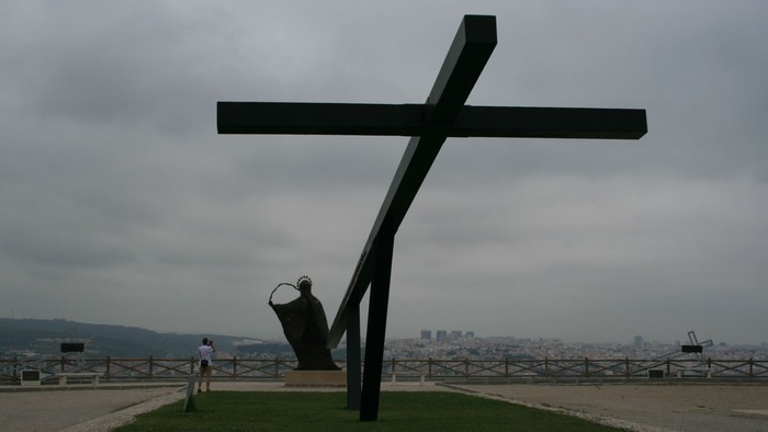 Ein großer schwarzer Kreuzmonument steht auf einem grasbewachsenen Hügel mit Stadtblick.