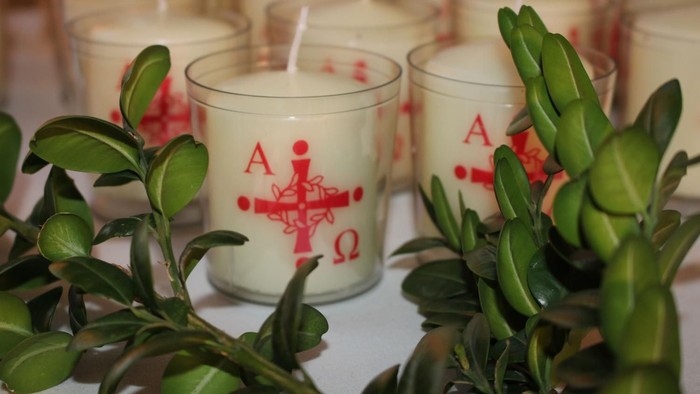 Weiße Kerzen mit rotem Kreuz und Buchstaben, umgeben von grünen Blättern