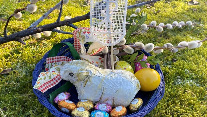 Ein Osternest mit bunten Eiern, einer weißen Osterhasenfigur und einer Zitrone auf einem blauen Korb im Gras