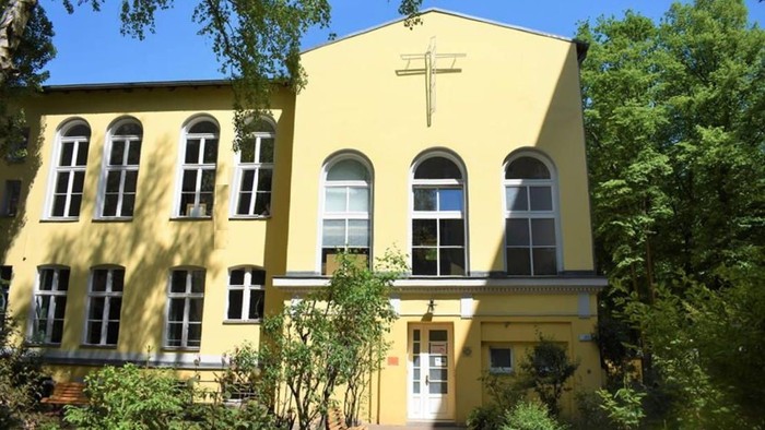 Helles gelbes Kirchengebäude mit Bogenfenstern und einem Kreuzemblem, umgeben von Grünflächen.