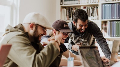 Ein Mann zeigt etwas auf einem Laptop, während zwei andere Personen interessiert zusehen. Im Hintergrund ein Bücherregal.