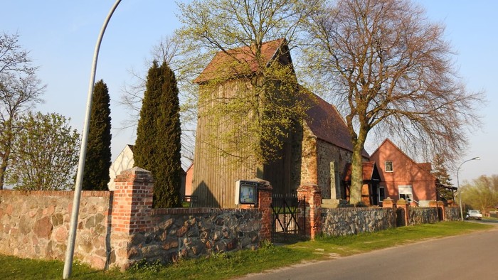 Ein rotes Backsteingebäude mit spitzem Dach und hohem Tor, umgeben von Bäumen und einem Laternenpfahl.