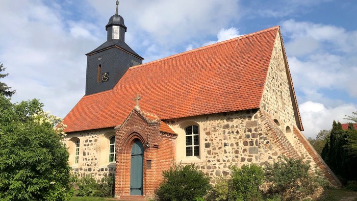 Kleine Kirche mit rotem Dach und Turm, umgeben von Grün.