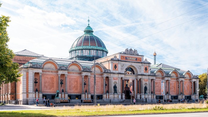 Stor og præget bygning med kuppel og bogstaver på fasaden, beliggende ved en vej med græsplæne foran