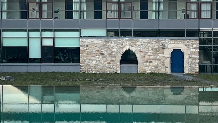 Modernes Gebäude mit reflektierender Glasfassade und gewölbtem Steineingang neben einer ruhigen Wasseroberfläche.