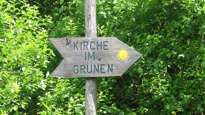 Hölzernes Wegweiser-Schild mit zwei Richtungsanzeigen, die auf „Kirche“ und „Grünen“ zeigen, umgeben von grünem Laub.