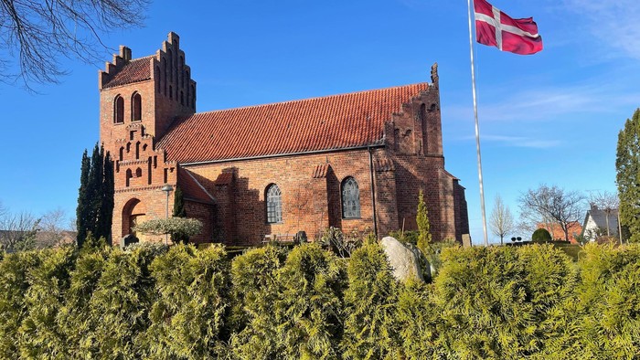 Kirke af røde mursten med tårn og dansk flag på en klar blå dag