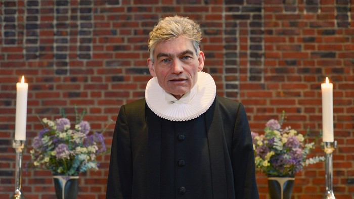 Mand i sort kappe og hvid krage står forud for mur med blomster og lygter