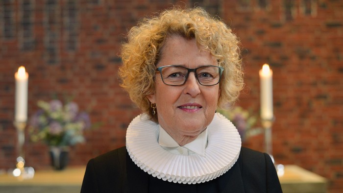 En kvinde med hårbånd og briller står foran murvæg med lysstake og blomster.