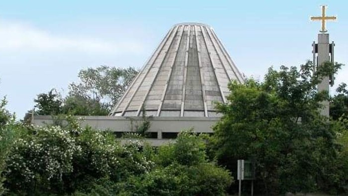 Das Bild zeigt eine moderne Kirche mit einem pyramidenförmigen Dach, umgeben von Bäumen und einem Kreuz.