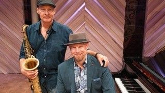 Zwei Männer mit Vintage-Hüten posieren zusammen, einer hält ein Saxophon neben einem Flügel.