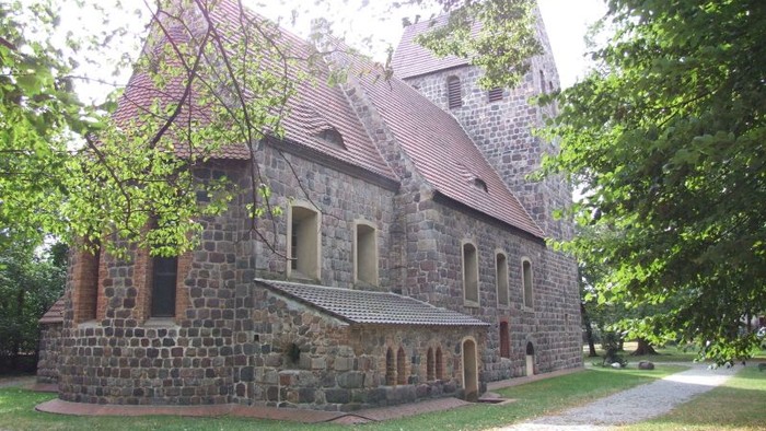 Altes steinernes Gebäude mit spitzem Dach und Turm, umgeben von Bäumen
