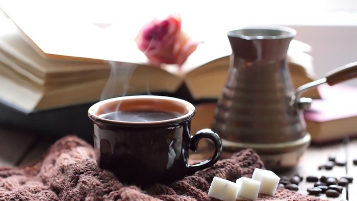 Ein schwarzer Becher mit Kaffee, Zuckerwürfel, Bücher und eine Rose auf einem Tisch
