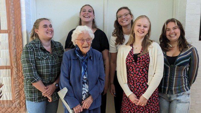 En gruppe af fem kvinder, heriblandt en ældre kvinde med stok, står sammen i et rum med klokke på væggen.