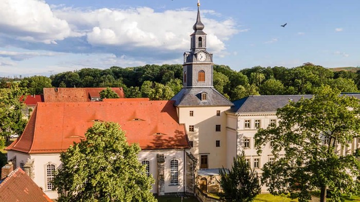 Ein weißes Gebäude mit rotem Dach und einem hohen Turm, umgeben von Bäumen und grünem Gras unter einem klaren blauen Himmel