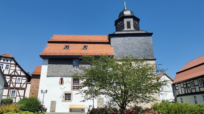 Das Bild zeigt eine Kirche mit einem Turm und rotem Dach, umgeben von grünen Büschen und Bäumen