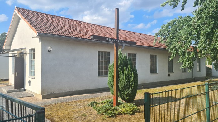 Kleines weißes Gebäude mit rotem Dach und Schornstein, umgeben von einem Zaun und Grünfläche