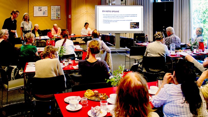 En gruppe af mennesker deltager i en workshop i et rum med bord og stoler, mens der vises en præsentation på en skærm.