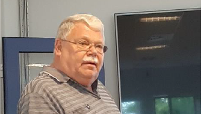 En ældre mand med hvidt hår og briller står foran en blå vegn.