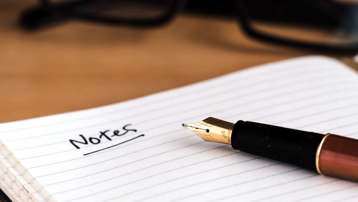 En notebog og en pen på en skrivebord med en par briller i baggrunden