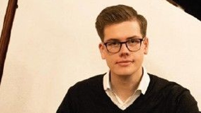 Ein junger Mann mit Brille lehnt an einem Geländer, darunter eine Frau mit Brille vor grünem Blattwerk