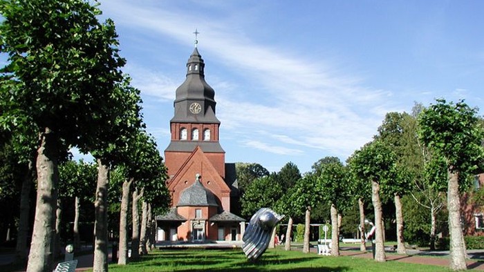 Kirchturm mit Uhr und Kreuz, umgeben von Bäumen und Park