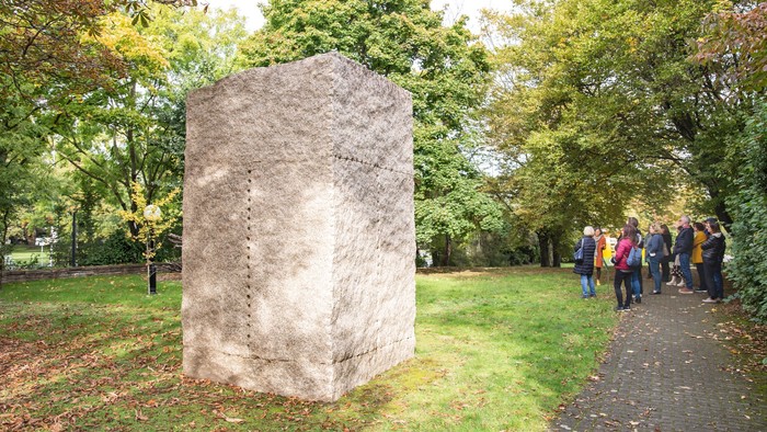 Ein großer Stein in einem Park, umgeben von Bäumen, und eine Gruppe von Menschen geht an ihm vorbei