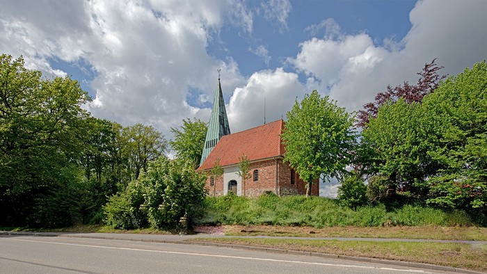 Kleine Kirche mit hohem Turm, umgeben von Bäumen und Büschen, an einer Straße