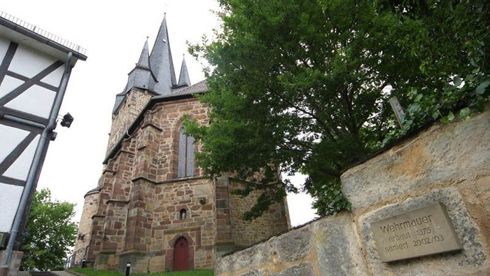 Steinerne Kirche mit hohen Türmen und einer roten Tür, umgeben von Bäumen und einer Steinmauer mit Gedenktafel.