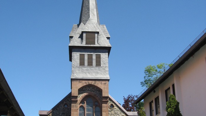 Kirche mit hohem Kirchturm und Kreuz, davor steht ein weißer Kleintransporter.