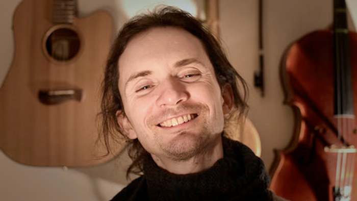 Ein Mann mit dunklem Haar und Bart lächelt vor Gitarren und einem Cello.