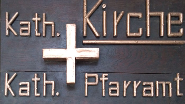 Das Bild zeigt ein hölzernes Schild mit den Namen Kath. Kirche und Kath. Pfarramt.