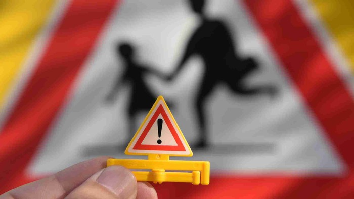 Hand hält gelbes Warndreieck mit Ausrufezeichen vor unscharfem Verkehrsschild mit Kindern.