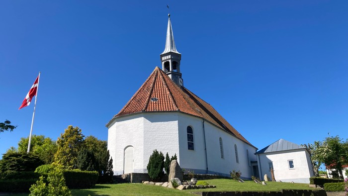 En hvid kirke med spiret og rødt tag, flagstang med danskirkeflag ved siden.