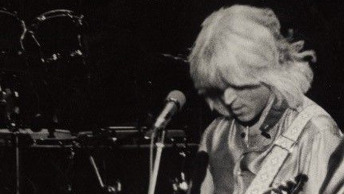 To guitarister på scene, en med blond hår og den anden med mørkt hår, begge spiller guitarer og synger i mikrofoner