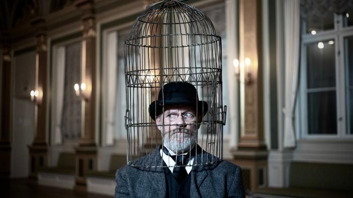 Mand med bur på hovedet, iført jakkesæt og hat, der står foran en bygning med store vinduer.