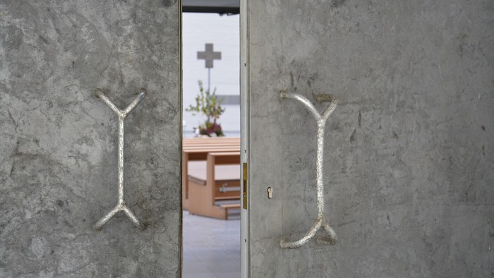 Zwei graue Türen mit Metallgriffen vor einem Raum mit Kreuz und Tisch