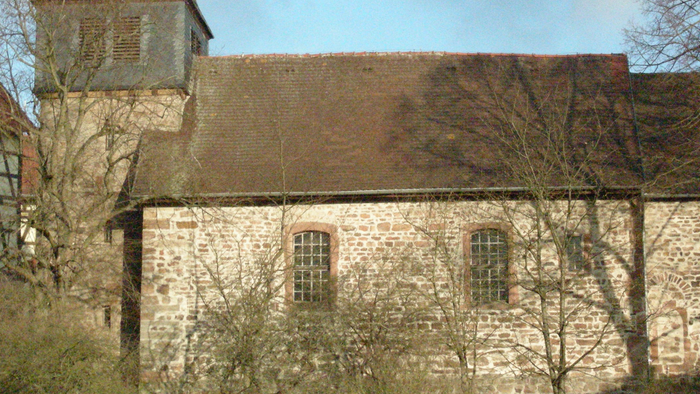 Alte Kirche mit hohem Turm und spitzem Dach, umgeben von Bäumen und Büschen