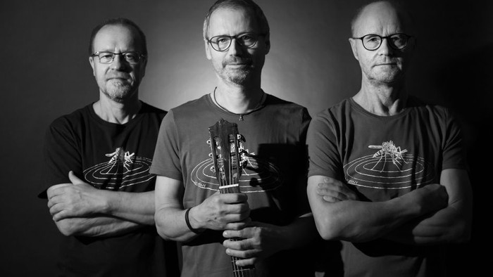 Drei Männer mit Brillen und T-Shirts, einer hält eine Gitarre