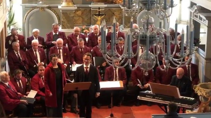 Ein Chor singt in einer Kirche, begleitet von einem Pianisten und einem Orchester.