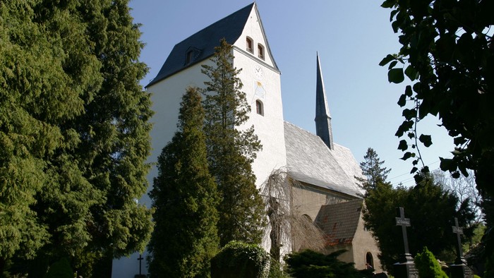 Weiße Kirche mit hohem Turm und spitzem Dach, umgeben von grünen Bäumen und Büschen