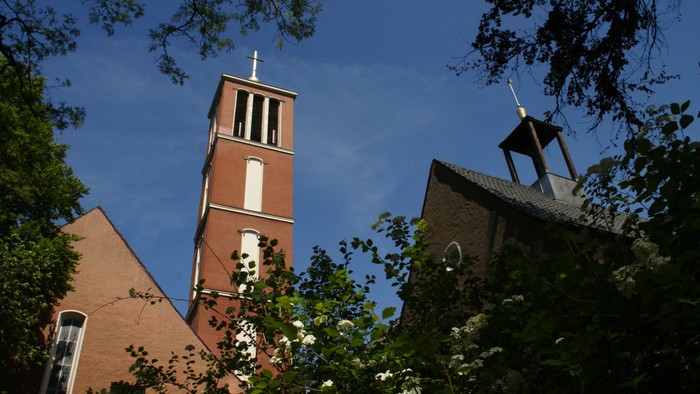 Ein roter Ziegelbau mit zwei Türmen und Kreuz, umgeben von Bäumen und Büschen