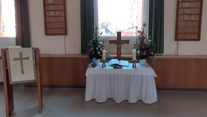 Ein Tisch mit weißer Tischdecke und Blumen in einem Raum mit zwei Kreuzen und Fenstern