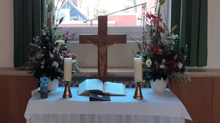 Ein Tisch mit weißer Tischdecke und Blumen in einem Raum mit zwei Kreuzen und Fenstern