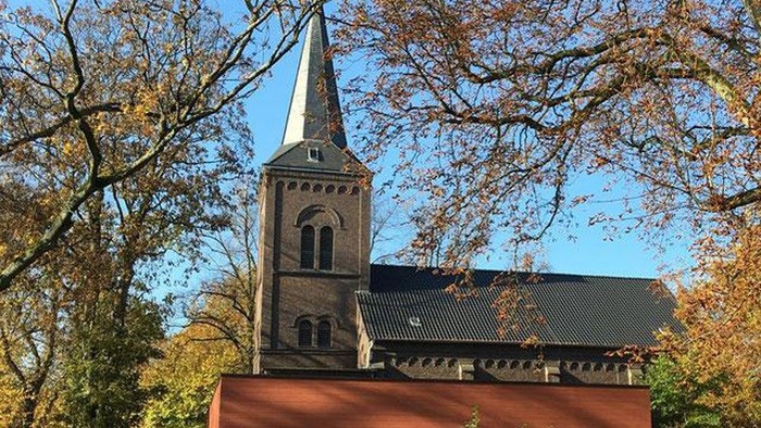 Ein Kirchengebäude mit hohem Turm und spitzen Dach, umgeben von Bäumen im Herbstlaub