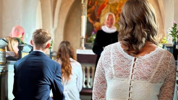 En bryllupsceremoni finder sted i en kirke med en præst og en gruppe gæster.