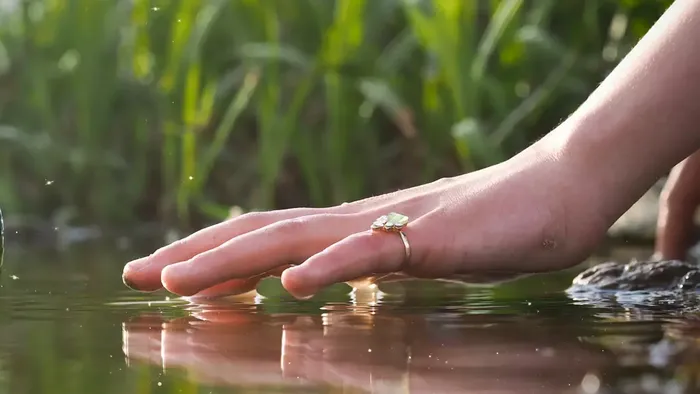 Eine Hand berührt sanft einen Stapel Kieselsteine im Wasser, umgeben von grünem Gras, mit der Aufschrift 