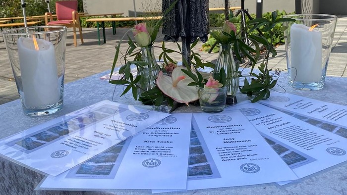 Auf einem Tisch stehen Kerzen, ein Kreuz und Blumen vor einem Gebäude mit Kreuz.