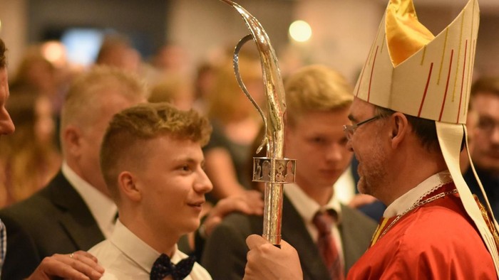 Ein Mann in einem roten Gewand und hohem Hut überreicht einem jungen Mann im Smoking einen Pokal.